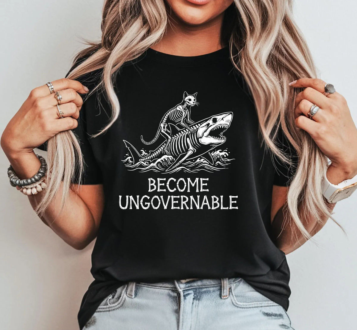 A woman wearing a black graphic t-shirt with a white print of a cat riding a shark and the text 'BECOME UNGOVERNABLE'.